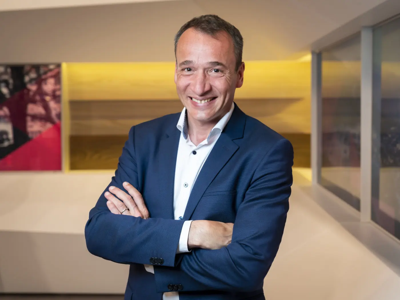 Uomo sorridente in giacca blu, con le braccia incrociate, in un ambiente moderno e luminoso.