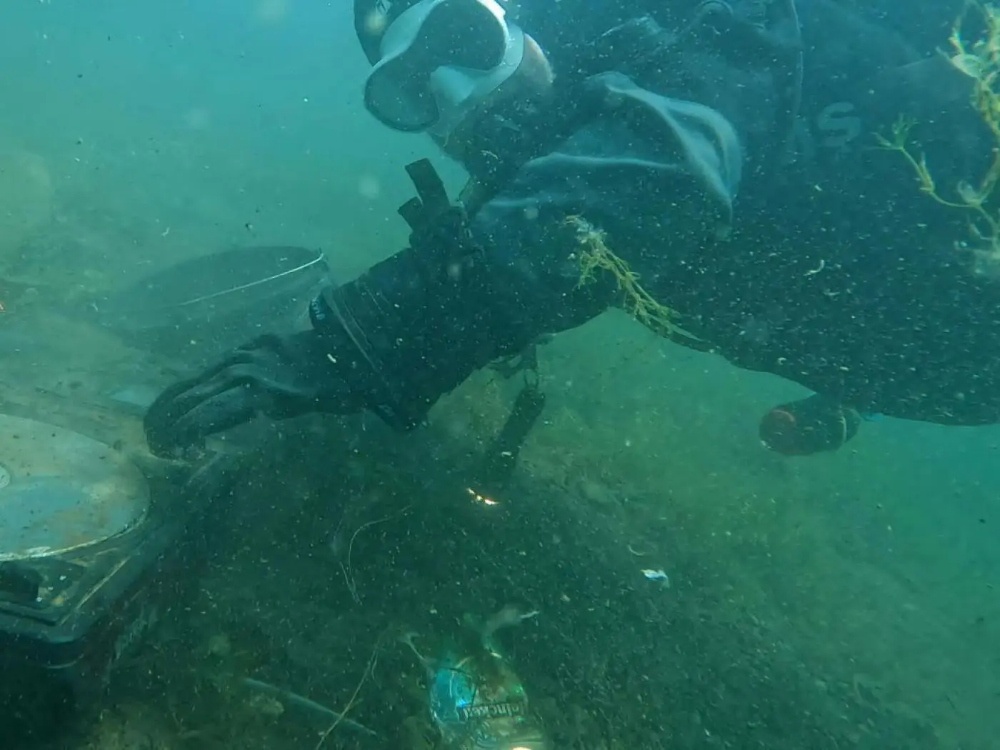 Un subacqueo esplora il fondale marino, toccando un oggetto metallico circondato da alghe. La visibilità è limitata e ci sono bolle d'aria nell'acqua.