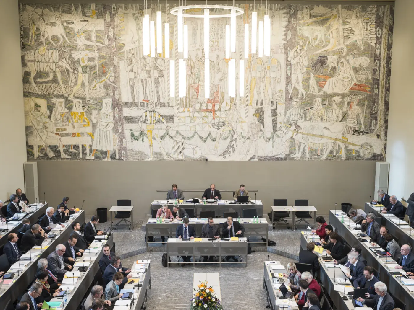 Una vista panoramica di una sala di dibattito con un grande murale sulla parete di fondo. Diversi membri siedono attorno a tavoli, alcuni parlano, mentre altri sono concentrati sui propri documenti. Al centro, un tavolo con dei fiori decorativi. L'illuminazione è moderna, con lampade a sospensione.