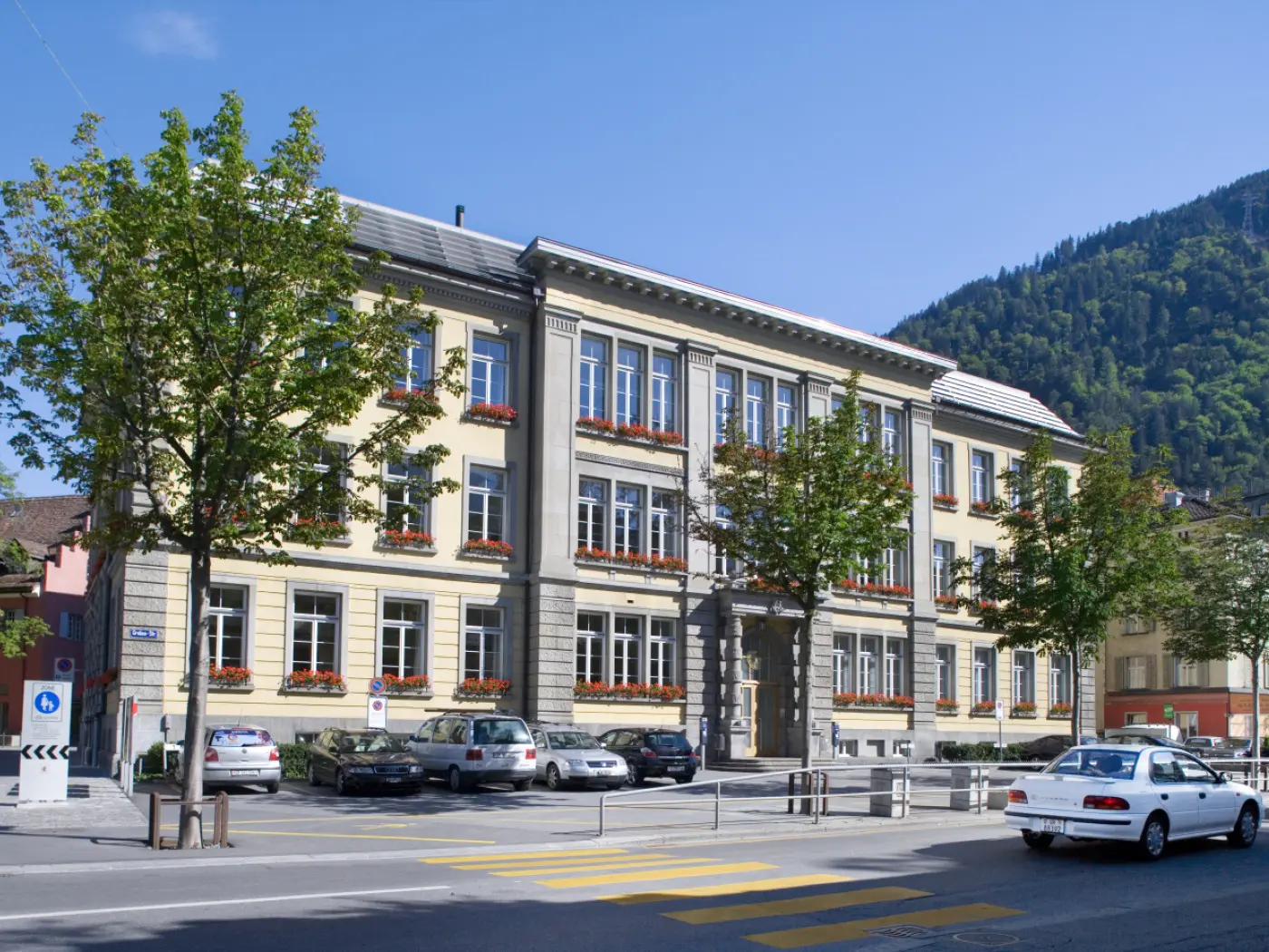 Edificio storico a tre piani con finestroni e fioriere, situato in una strada tranquilla. Sullo sfondo, una collina verde. Auto parcheggiate di fronte all'edificio.