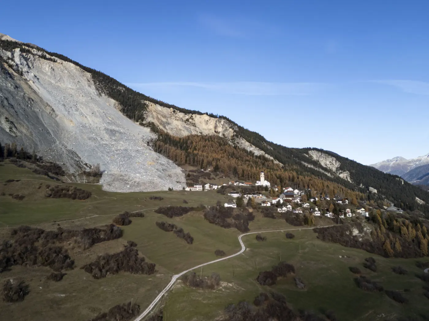 Un paesaggio montano con un villaggio pittoresco in basso. Sullo sfondo, una grande frana su una parete di roccia. Gli alberi dorati in autunno si stagliano contro il cielo blu. Un sentiero curvo attraversa il prato verde.