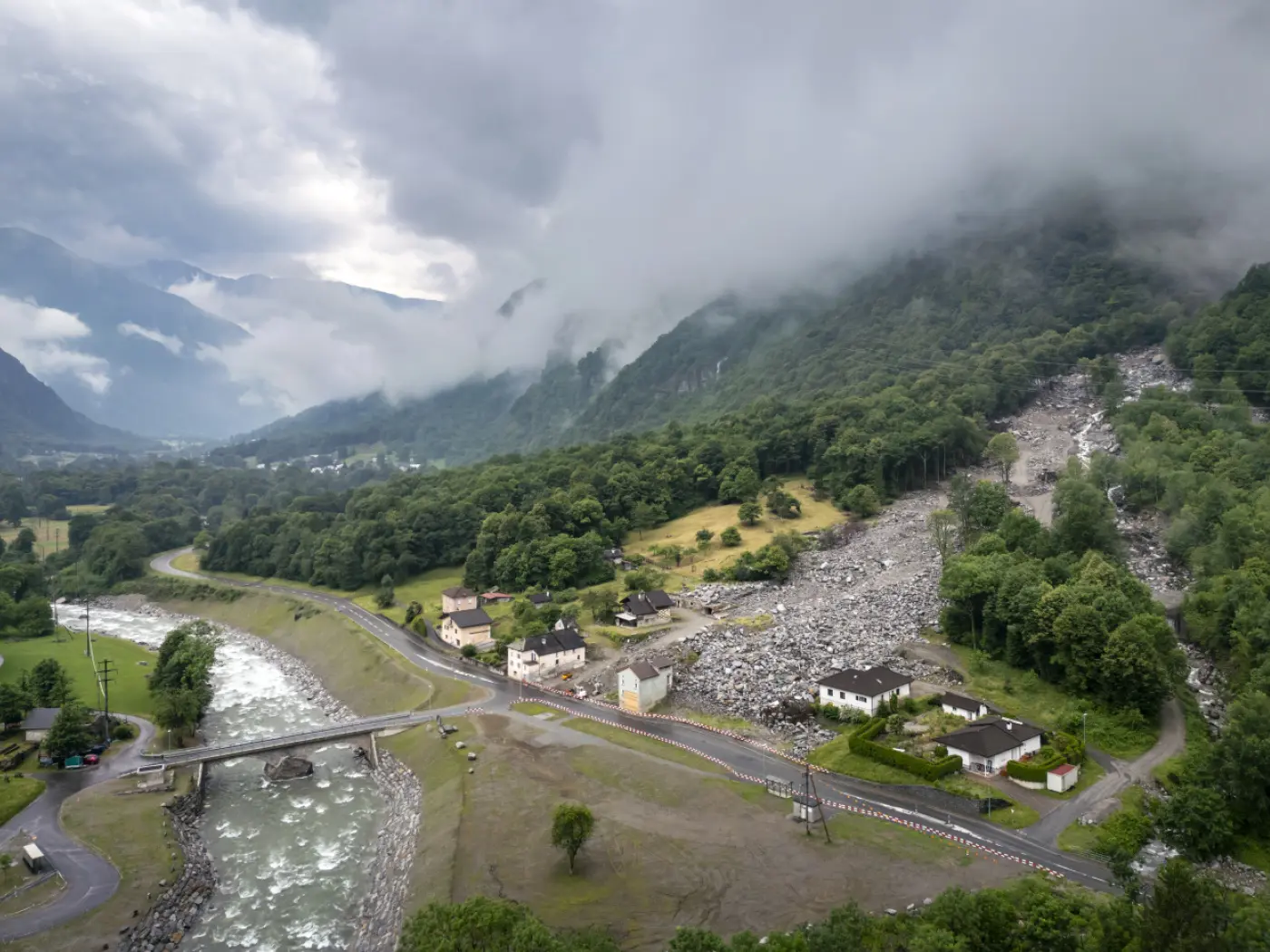 Frane che scendono da una montagna verde, con case danneggiate e un fiume che scorre vicino. Nubi basse coprono il cielo sopra il paesaggio colpito.