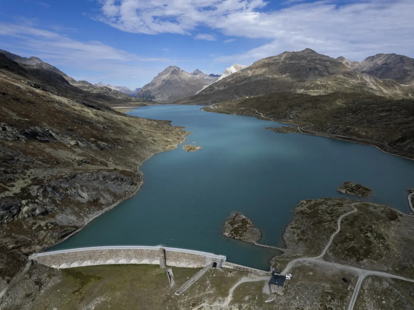 Panorama di un lago circondato da montagne, con una diga visibile in primo piano. Il cielo è parzialmente nuvoloso.