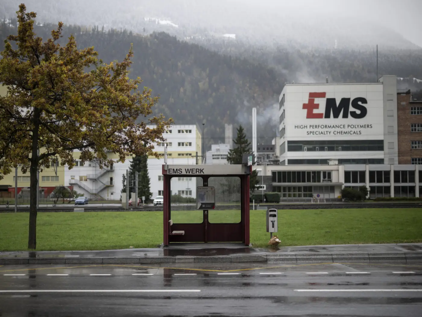 Deposito fermata dell’autobus con logo “EMS” in un’area industriale. Sullo sfondo si scorgono edifici moderni e colline coperte di nebbia. Alberi gialli ai lati della strada bagnata dalla pioggia.