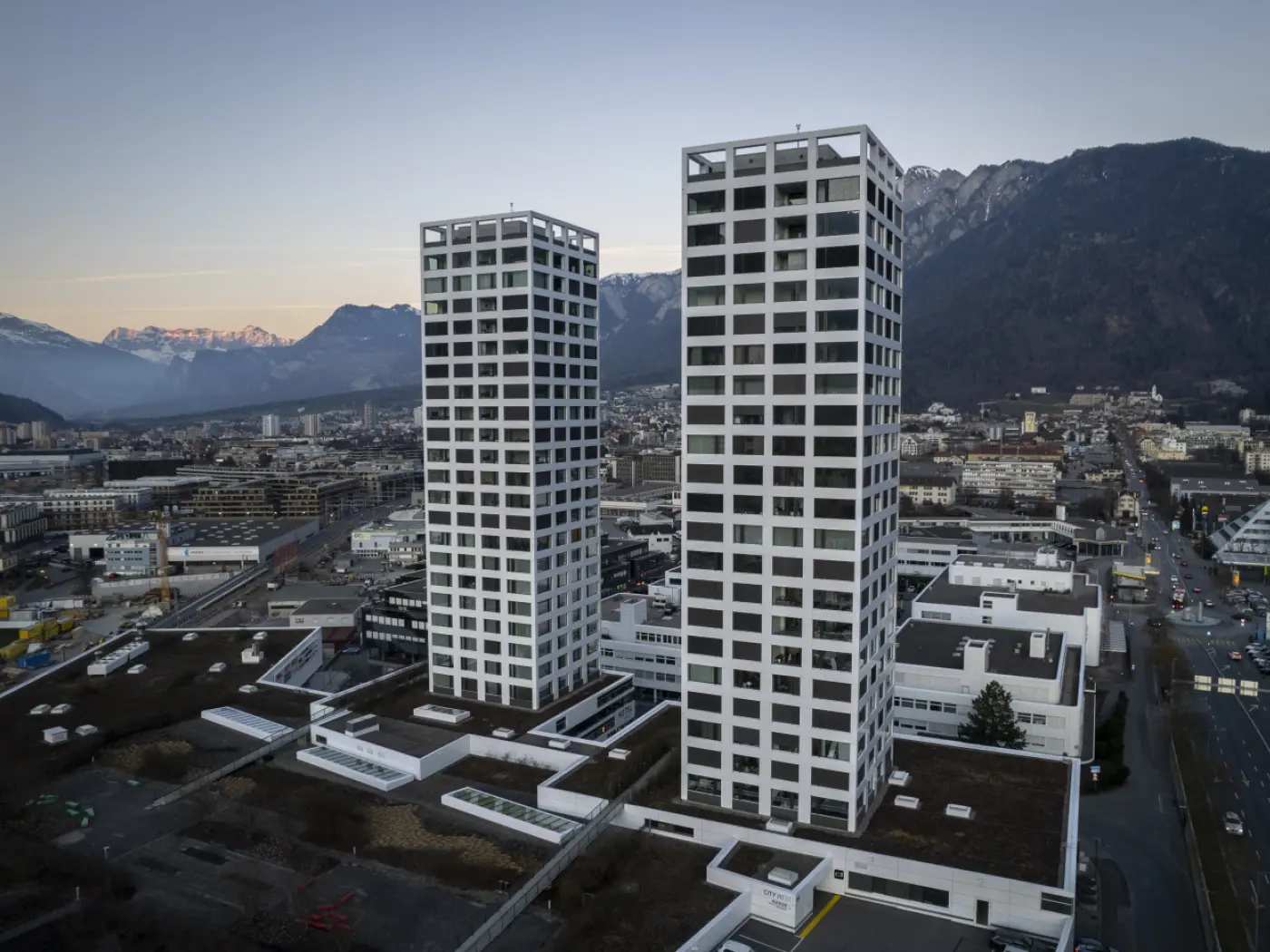 Due torri moderne di cemento bianco si ergono su un'area urbana, circondate da montagne e un paesaggio cittadino. Il cielo è sereno e si intravedono strade e edifici circostanti.