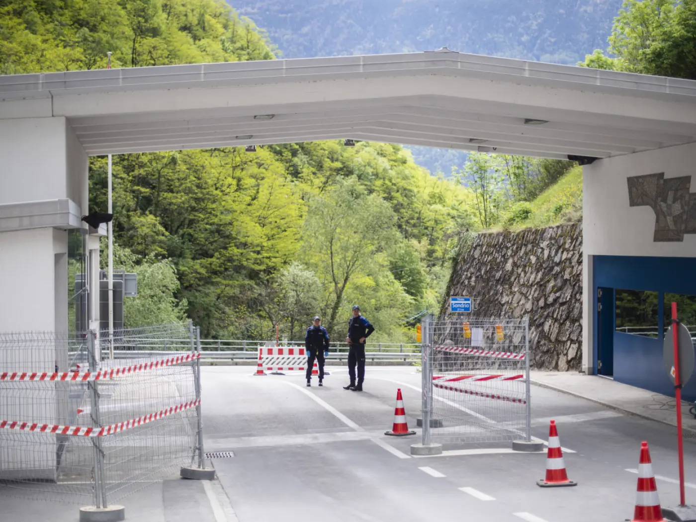 Settore di controllo con barriere di sicurezza, due agenti di polizia in uniforme vigilano all'ingresso. Sullo sfondo si vedono alberi verdi e montagne.