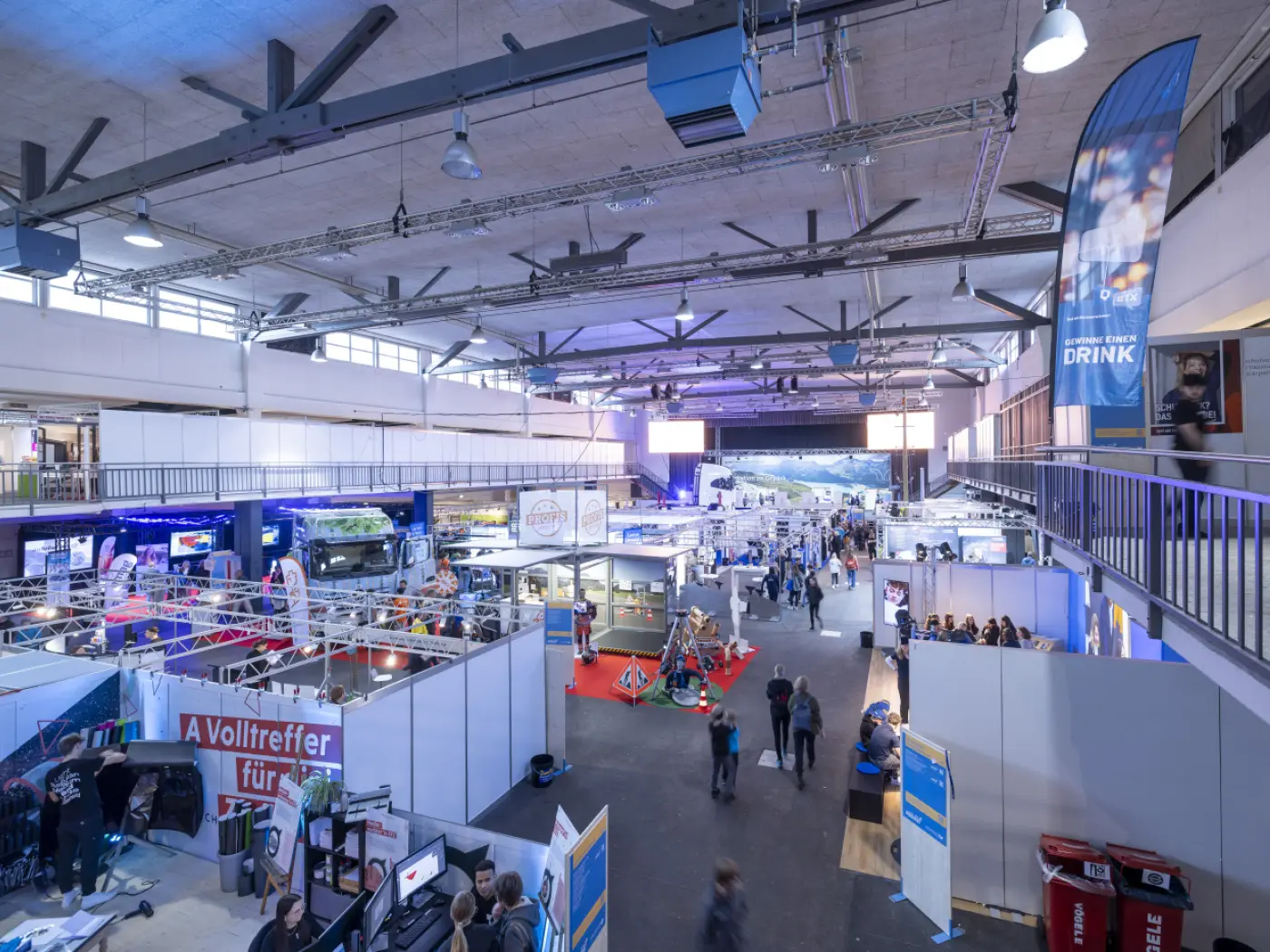 Una vista panoramica di una fiera o un evento espositivo, con vari stand e visitatori che si muovono tra le aree espositive. Strutture e manifesti decorano lo spazio, creando un'atmosfera vivace.