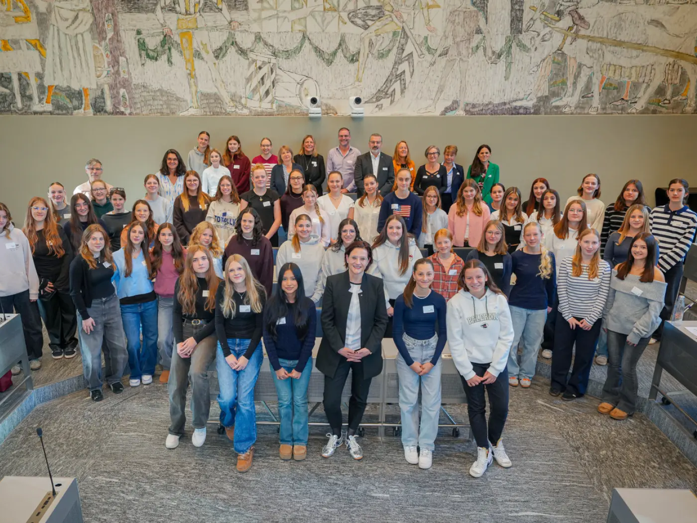 Gruppo di persone sorridenti in una sala con una grande opera murale sullo sfondo. Ci sono giovani donne e uomini di diverse età, alcuni indossano etichette con il nome. La scena trasmette un senso di comunità e collaborazione.