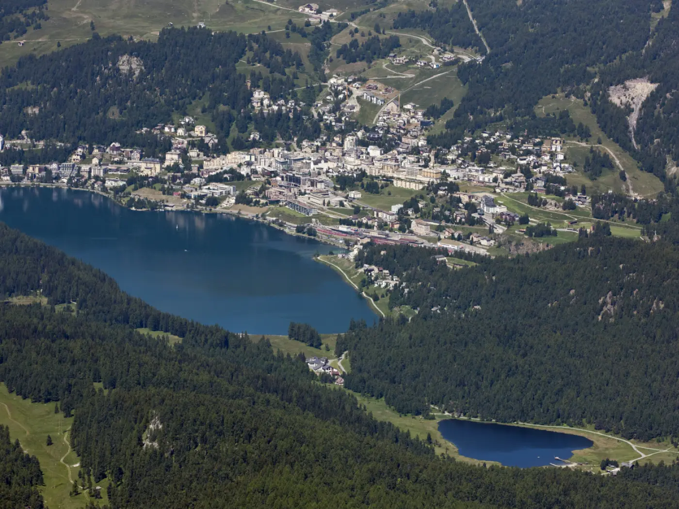 Vista panoramica di un lago circondato da montagne e una città. La vegetazione verde si estende lungo il lago, mentre le case sono sparse sulle colline. Il cielo è sereno.