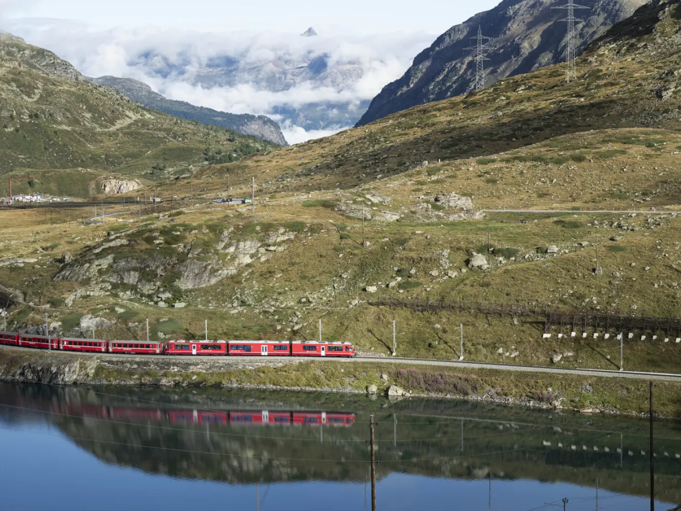 Un treno rosso percorre una pista ferroviaria lungo un lago in montagna, circondato da colline verdi e nuvole basse. Le cime montuose si stagliano sullo sfondo, riflettendosi nell'acqua calma.