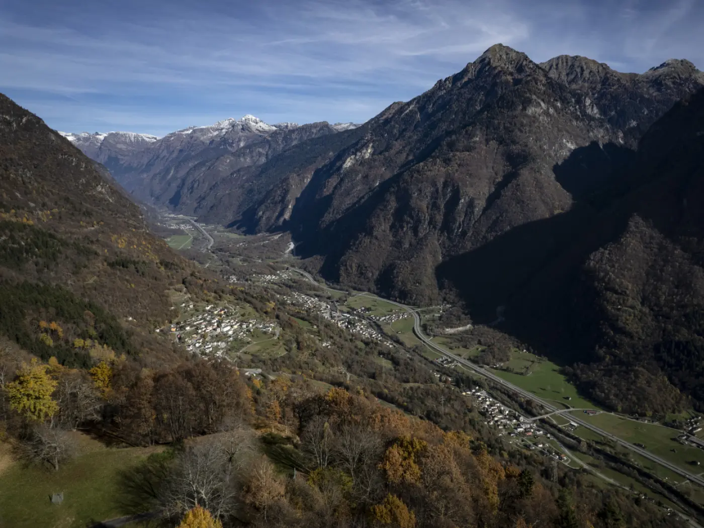 Vista panoramica di una valle montuosa con un fiume che scorre, circondata da alte montagne e boschi autunnali. La luce del sole illumina le cime innevate e il paesaggio sottostante.
