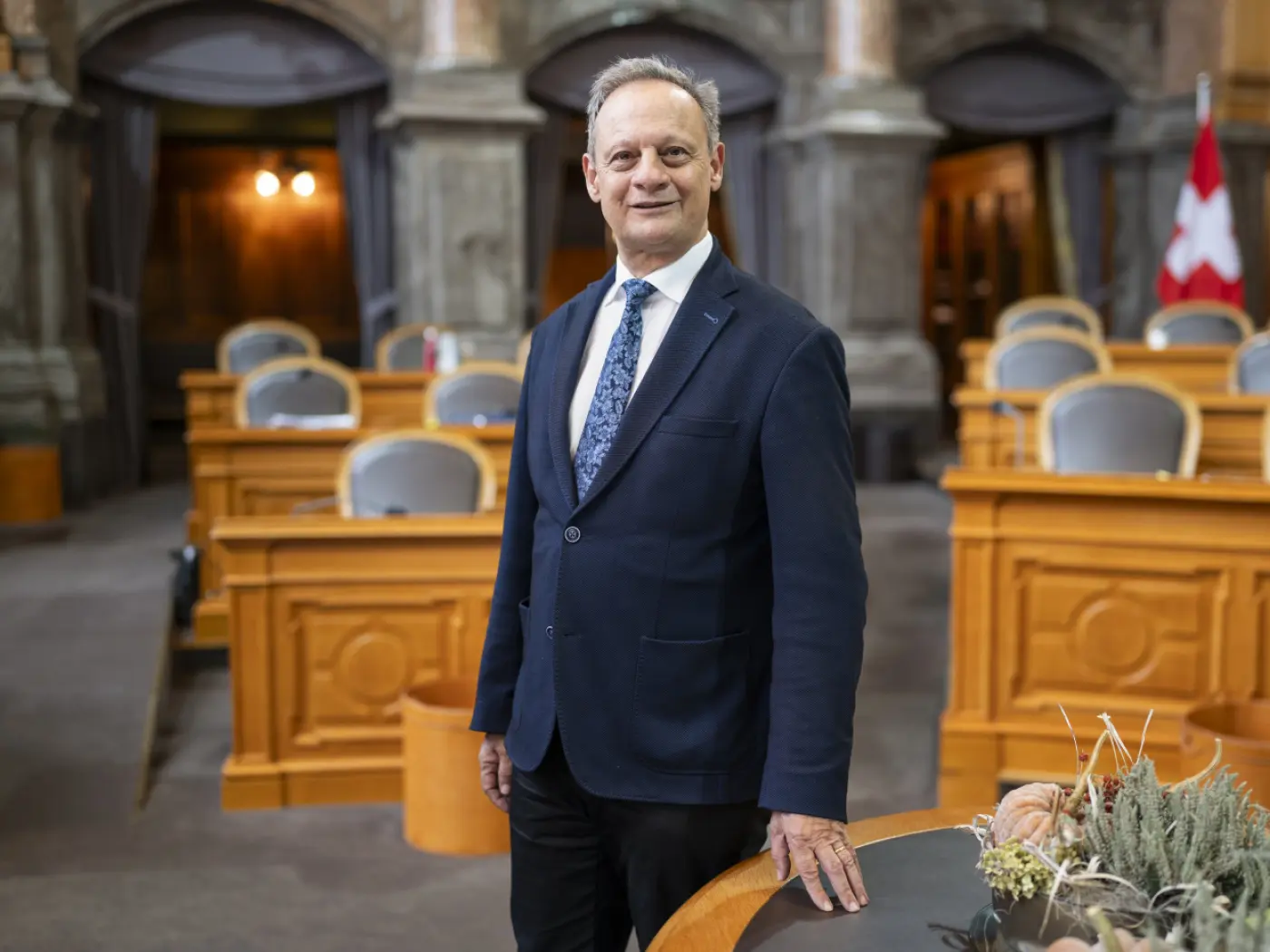 Un uomo in abito scuro e cravatta si trova in una sala legislativa. Sullo sfondo, banchi di legno e bandiera svizzera. L'atmosfera è formale e istituzionale.