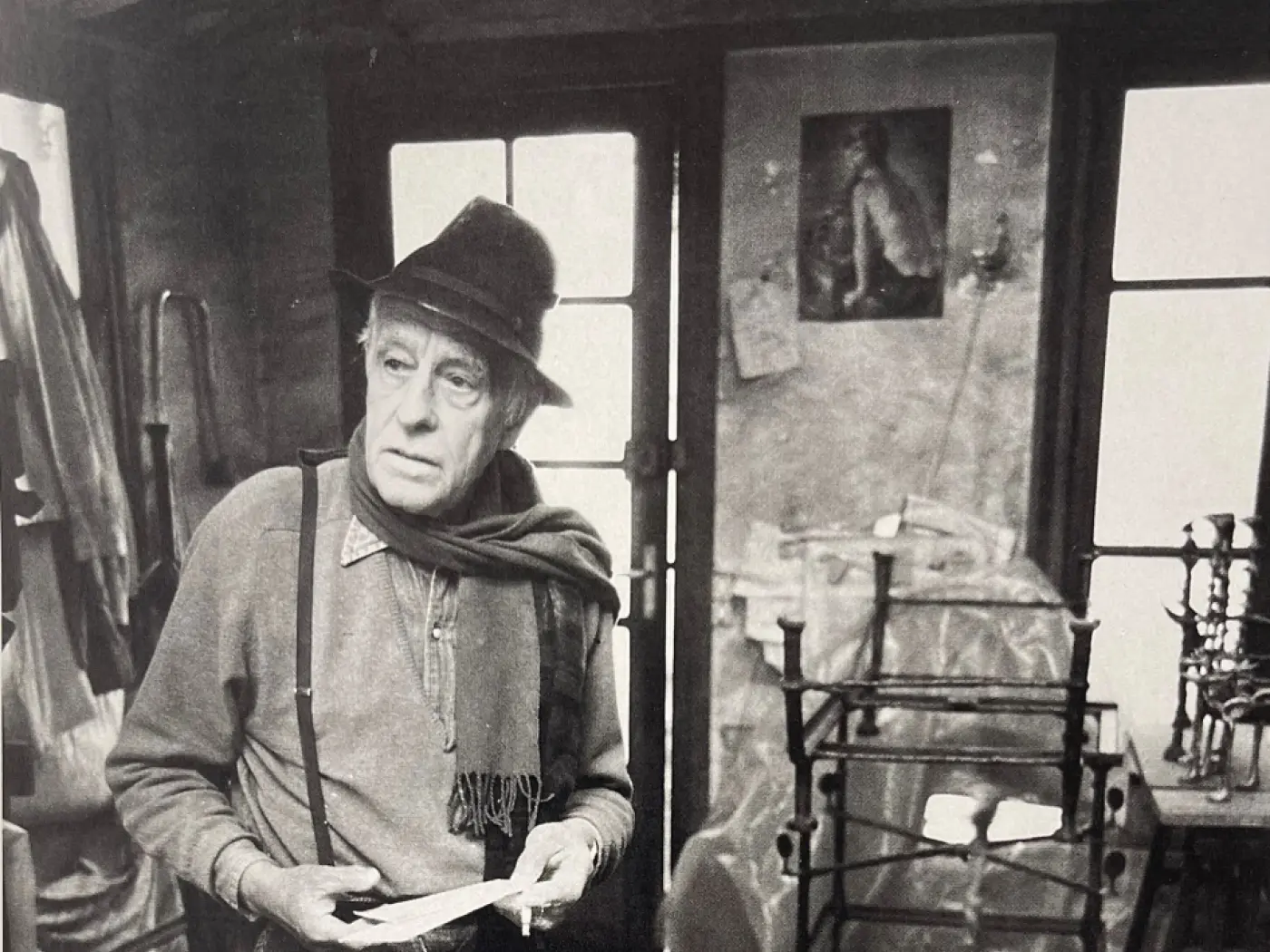 Un uomo anziano con un cappello e una sciarpa, in piedi in uno studio spoglio. Si guarda attorno, con espressione contemplativa, mentre tiene in mano un fog