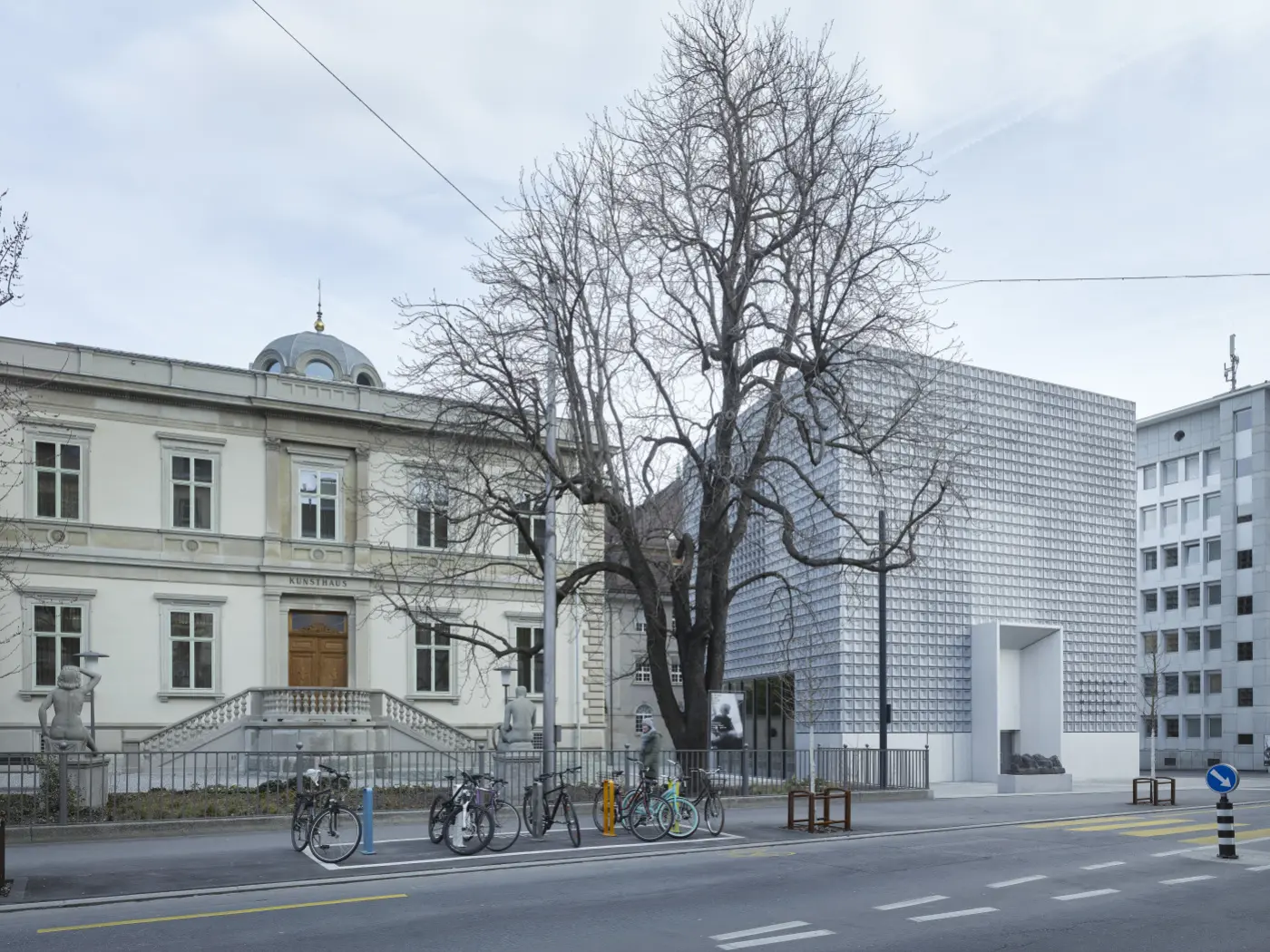 Una strada con un albero spoglio e due edifici. A sinistra, un edificio storico con una facciata ornata e porte in legno. A destra, una struttura moderna