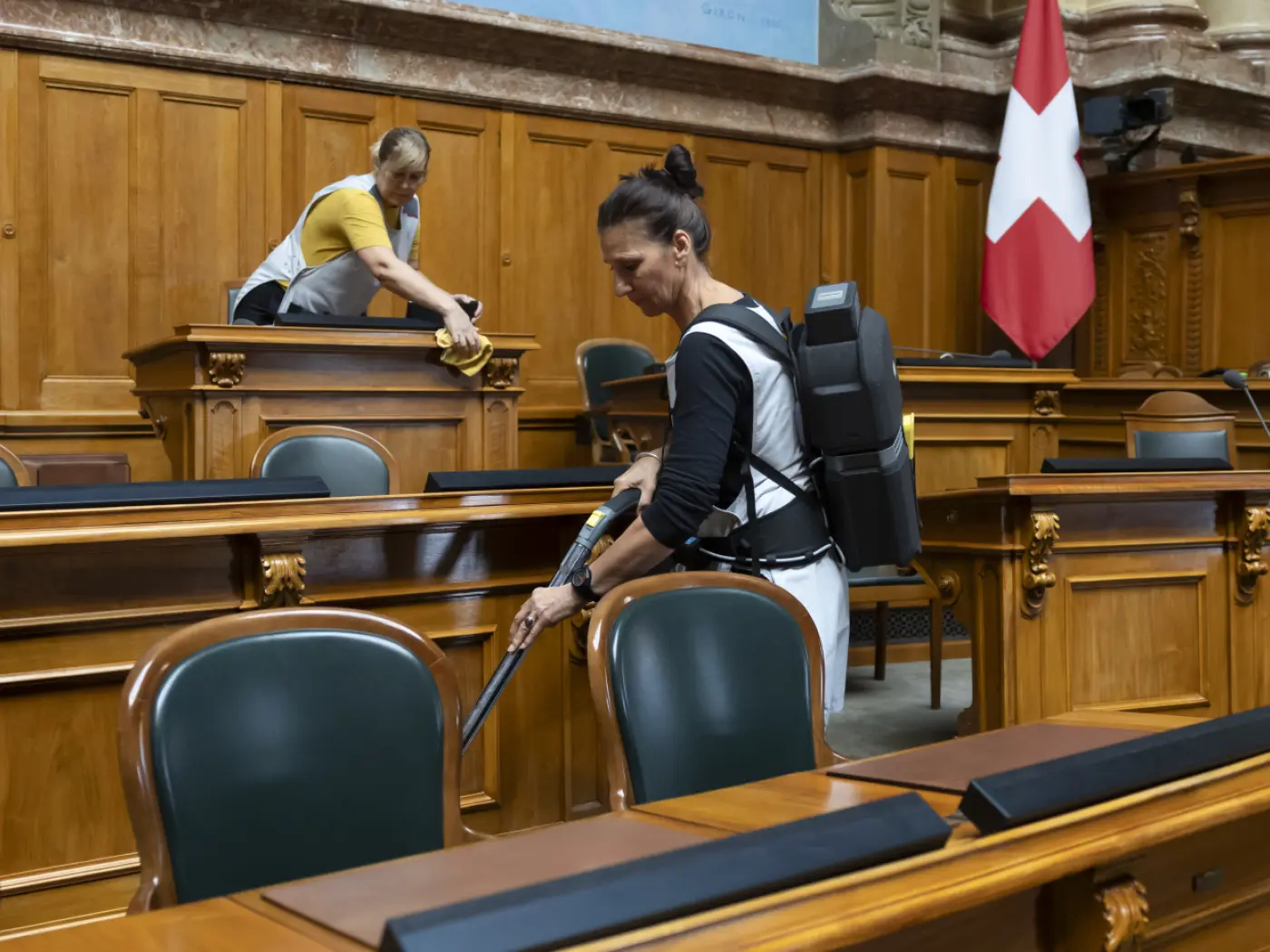 Due persone puliscono una sala di un edificio governativo. Una donna con uno zaino aspirapolvere pulisce i banchi, mentre un uomo spolvera. È visibile la band