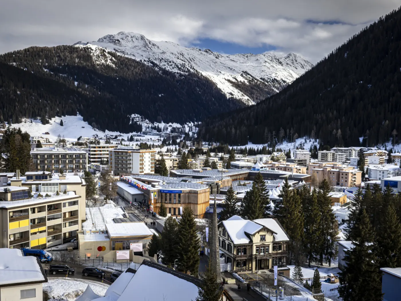 Panorama di una località montana coperta di neve, con edifici moderni e tradizionali, circondata da alberi e imponenti montagne innevate sullo sfondo