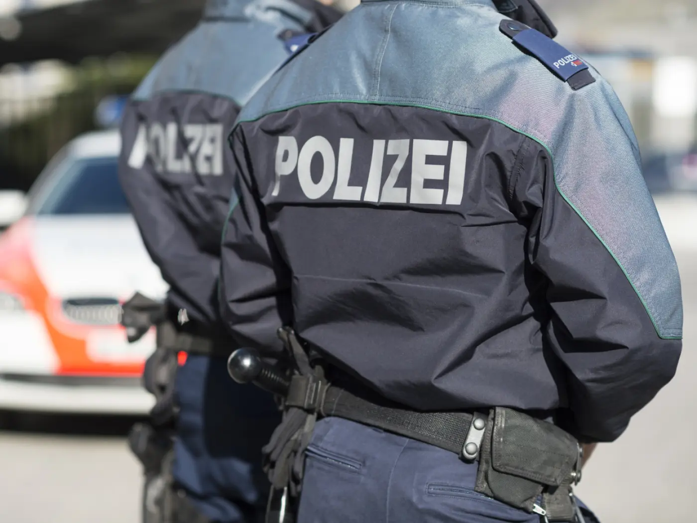 Due agenti di polizia in uniforme, con la scritta "POLIZEI" sul retro, in piedi di spalle. Un'auto della polizia è visibile sullo sfondo