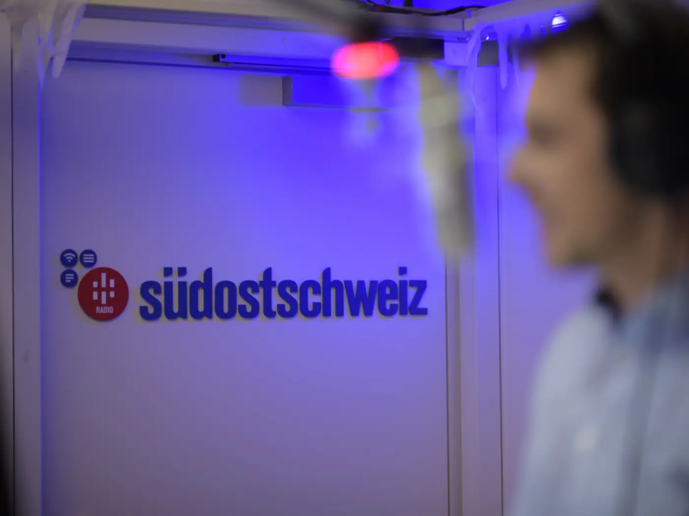 Sfondo con il logo di "südostschweiz radio" illuminato in blu, con una figura sfocata a destra che sembra essere una persona in un ambiente radiofonico