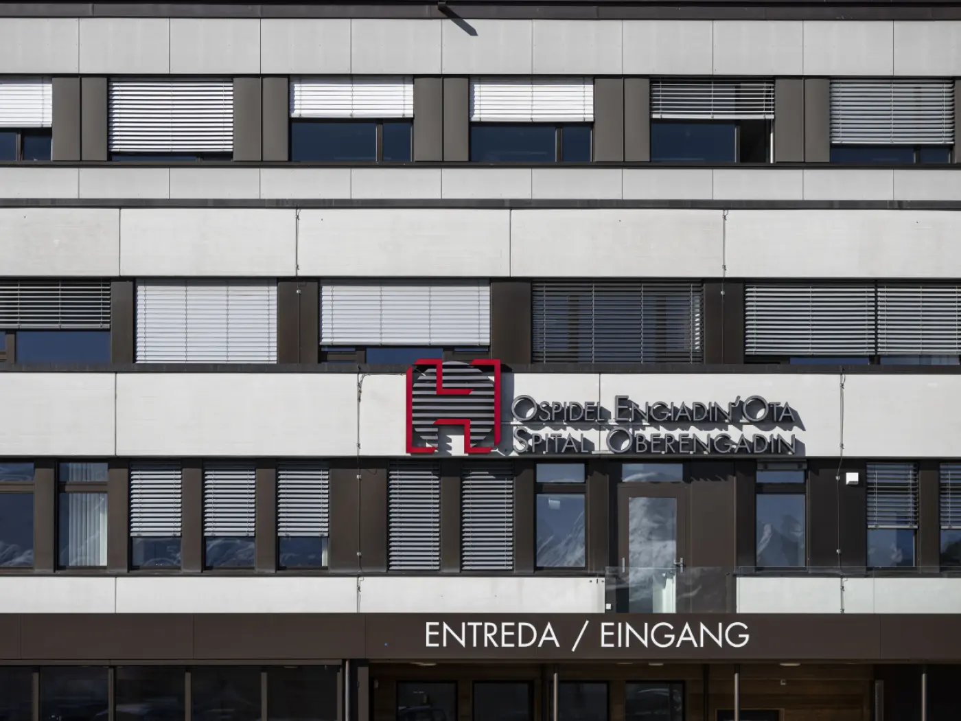 Facciata di un ospedale con il nome "Ospidel Engiadina" e la scritta "Entrata / Eingang" in diverse lingue. Le finestre sono dotate