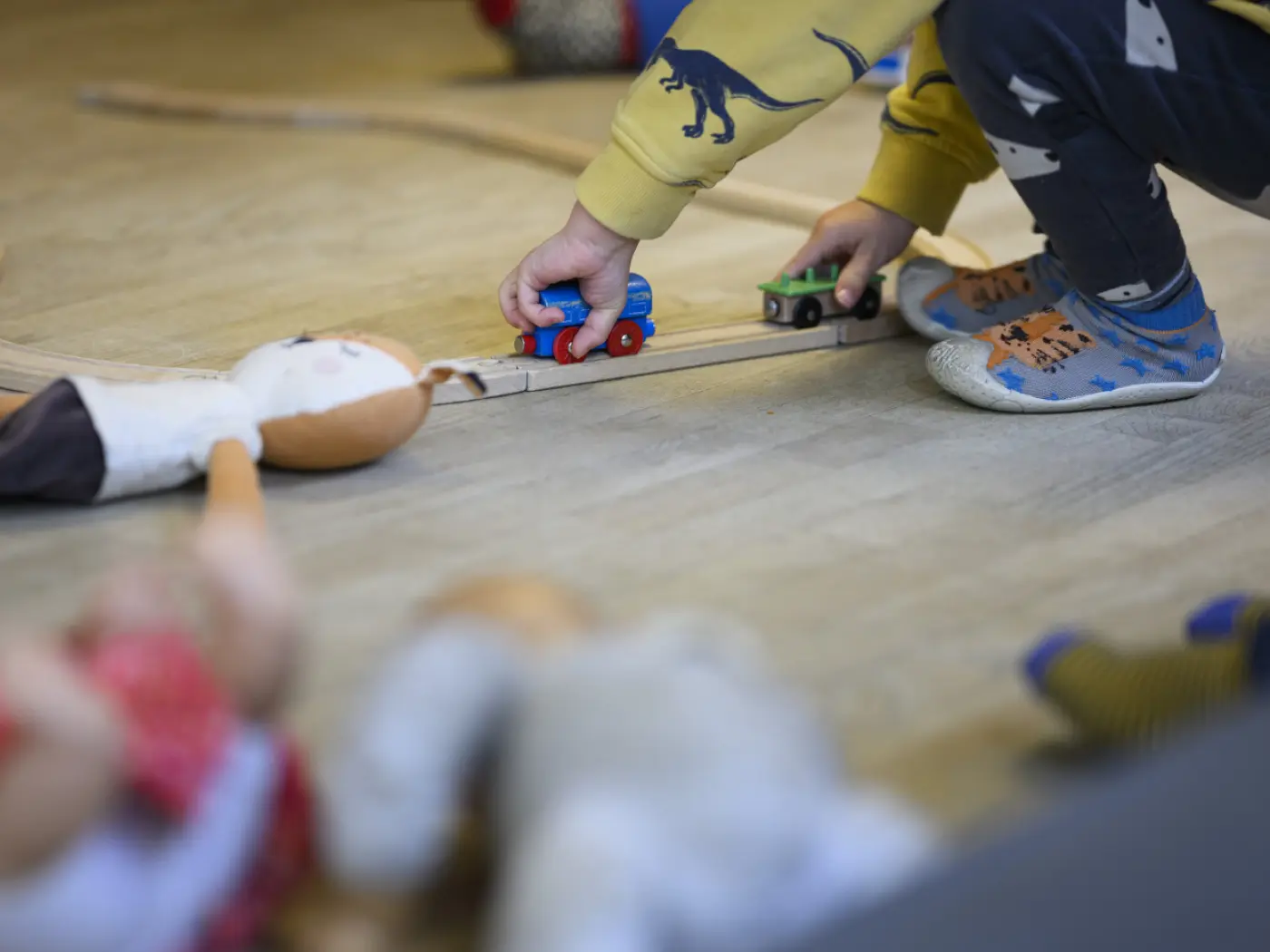 Un bambino gioca posizionando un trenino su un binario di legno. Intorno a lui ci sono alcune bambole e giocattoli sparsi sul pavimento.