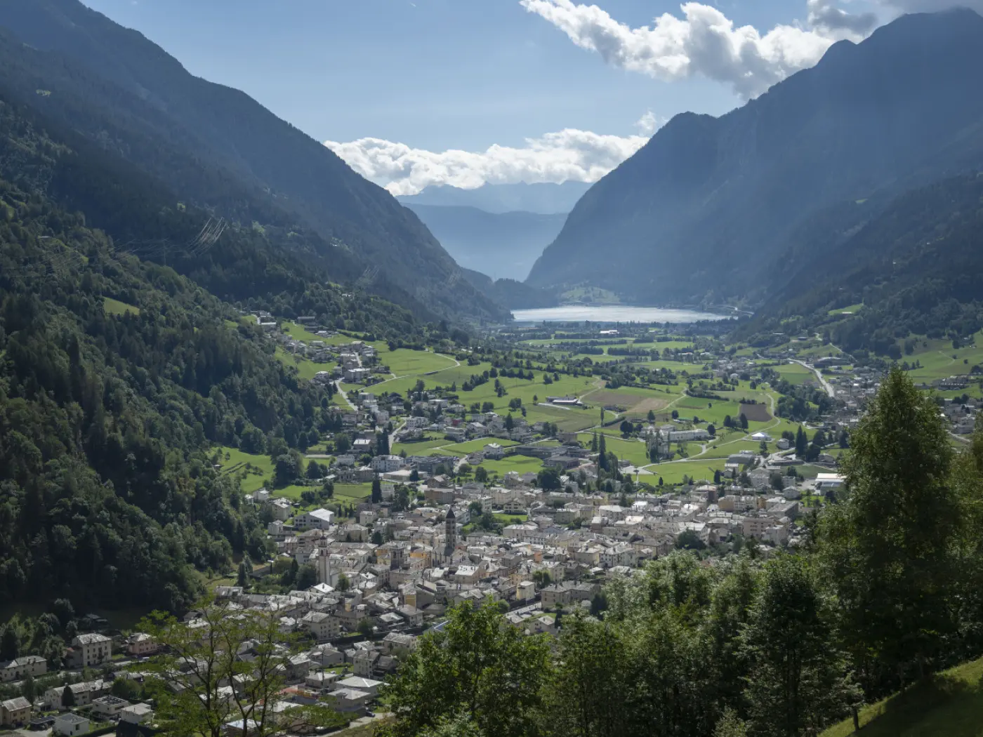 Veduta panoramica di una valle montana con un villaggio tra le colline verdi e le montagne. In lontananza si intravede un lago, mentre il cielo è sereno