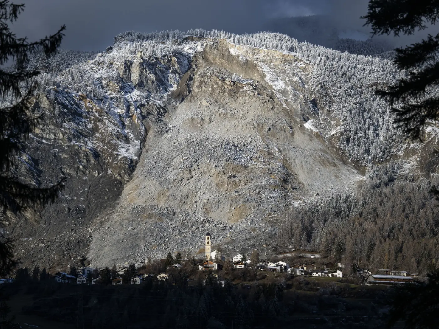 Una montagna innevata con una frana visibile e un piccolo villaggio sotto, caratterizzato da una chiesa con campanile. Gli alberi firmano il bordo dell'immagine