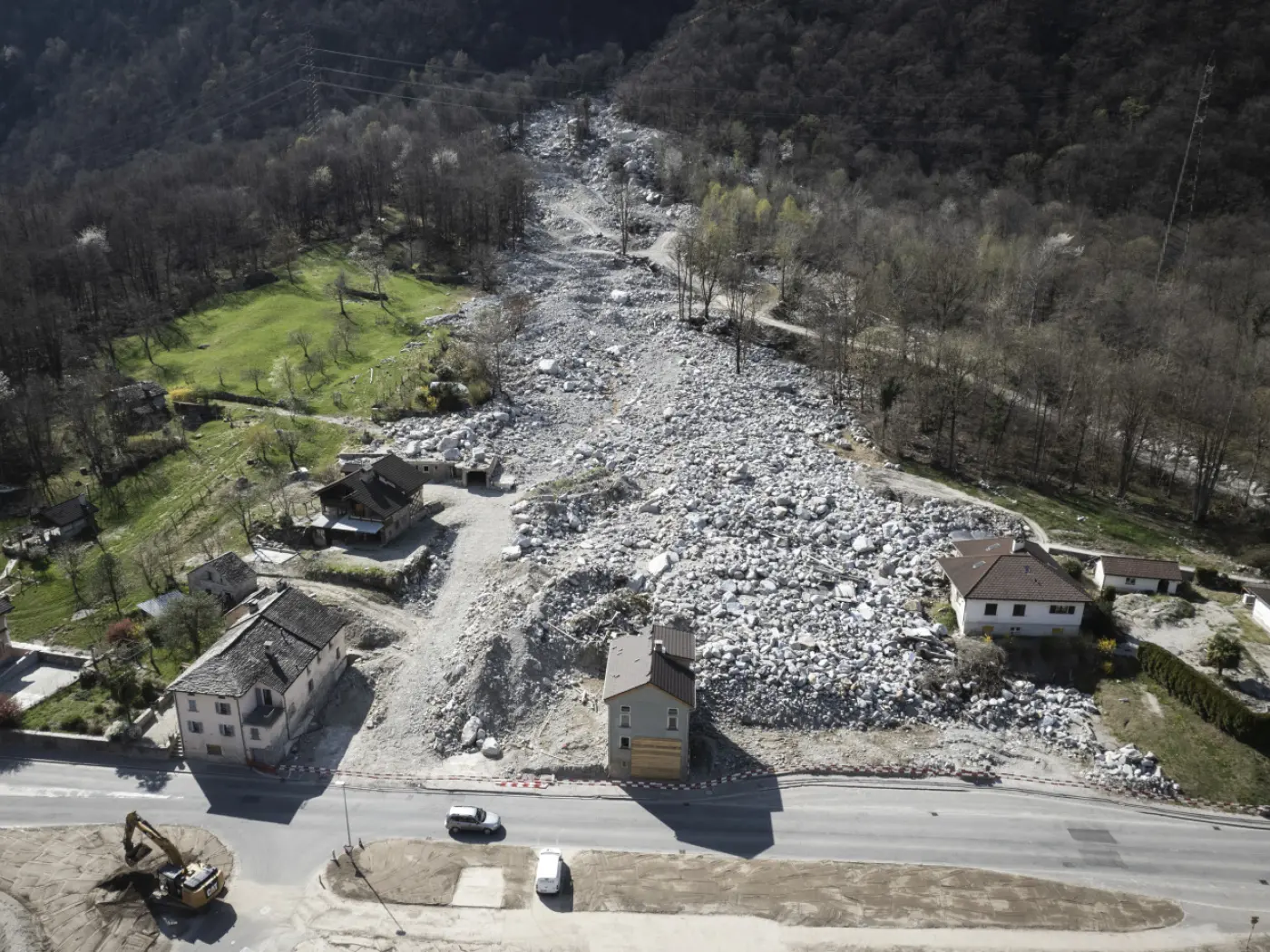 Paesaggio collinare devastato da un frana. Rocce e detriti coprono gran parte del terreno, mentre alcune case si trovano in prossimità della strada sottostante.