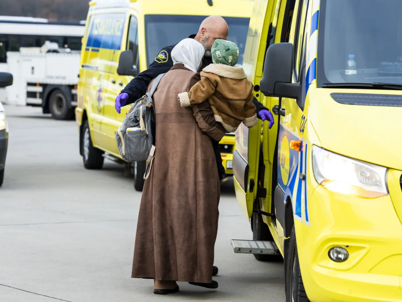 Una donna con un bambino in braccio, vestita con un cappotto marrone e un cappello, si avvicina a un'ambulanza gialla mentre un operatore sanitario la ai