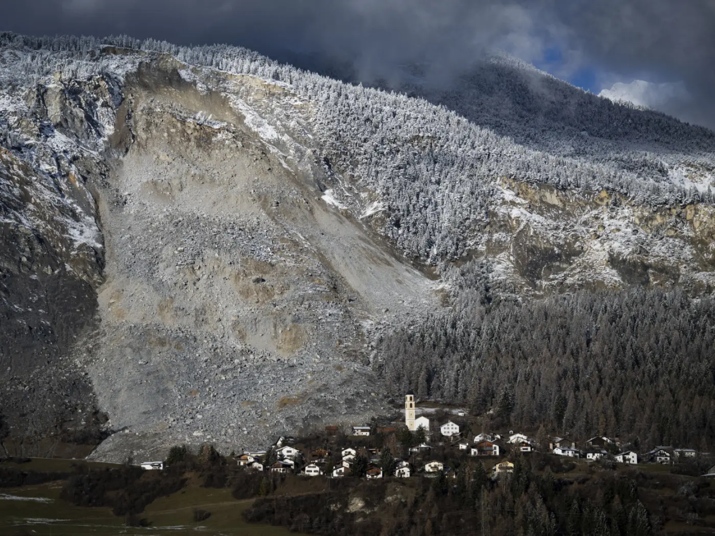 Un villaggio montano con case bianche in primo piano, circondato da alberi e montagne innevate. Una frana si è staccata sul lato della montagna, vis